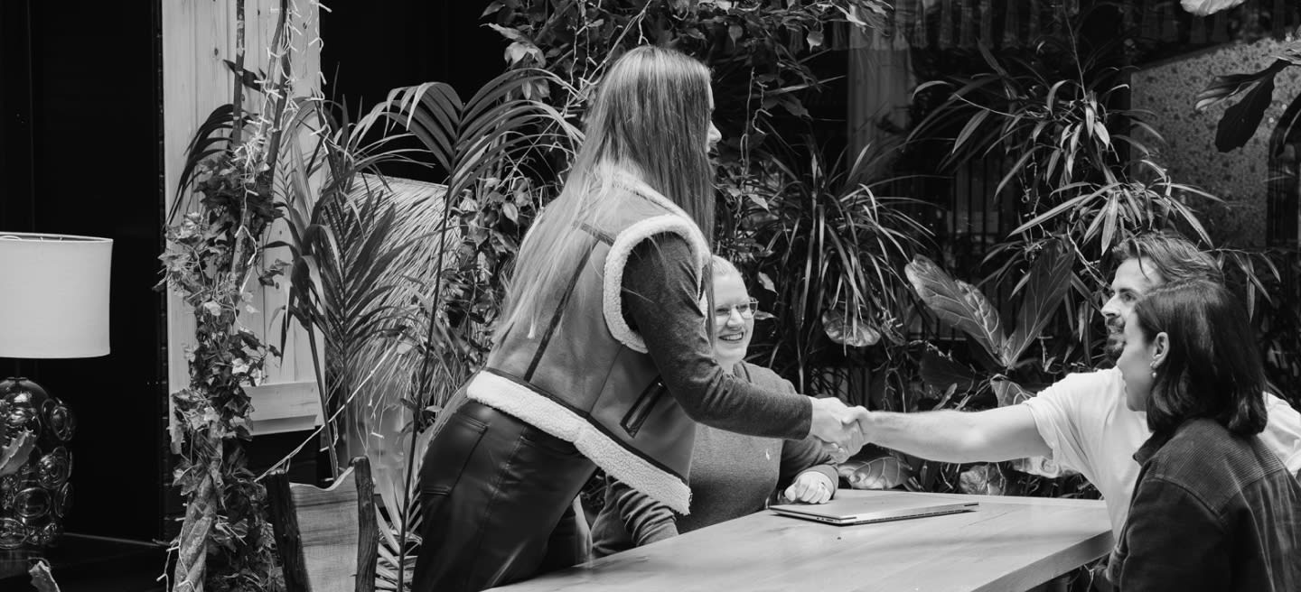 Kvinna skakar hand med man medan kollega sitter runt ett bord