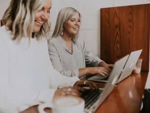 zwei blonde Frauen arbeiten lachend an Laptops