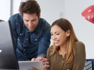 Two employees, one sitting at a desk and the other standing, working together on a laptop