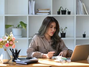 A person working from home, sitting at a desk with a laptop and holding a pen