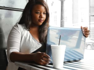 A young woman reading from her laptop as she sits at a table by the window of a diner.