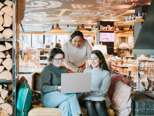 A cheerful group of three people, at a restaurant, one standing and two of them sittin on a couch, using a laptop