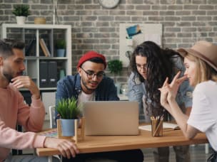 Four people collaborating at office table with laptop, notebooks, and plants