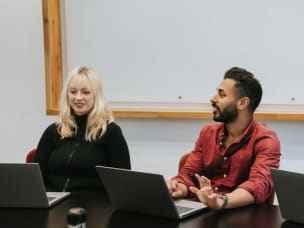 Two Awin employees sitting at a desk in a meeting room, engaging in a discussion while using their laptops.