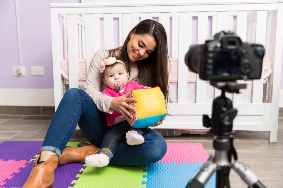 Person with child on lap holding toy block, with camera setup in front and crib in background