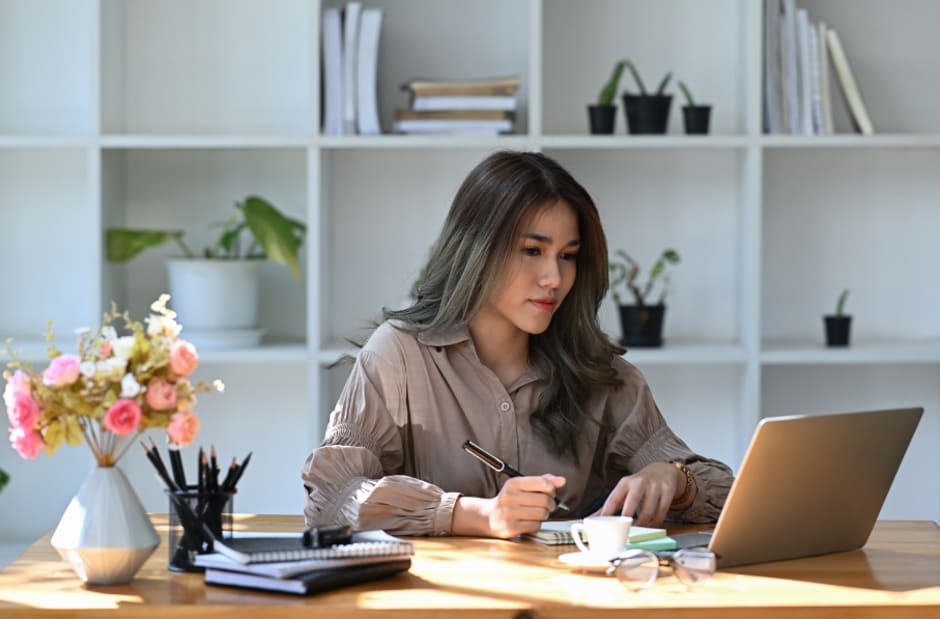 A person working from home, sitting at a desk with a laptop and holding a pen