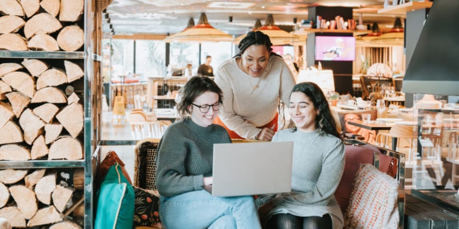 A cheerful group of three people, at a restaurant, one standing and two of them sittin on a couch, using a laptop
