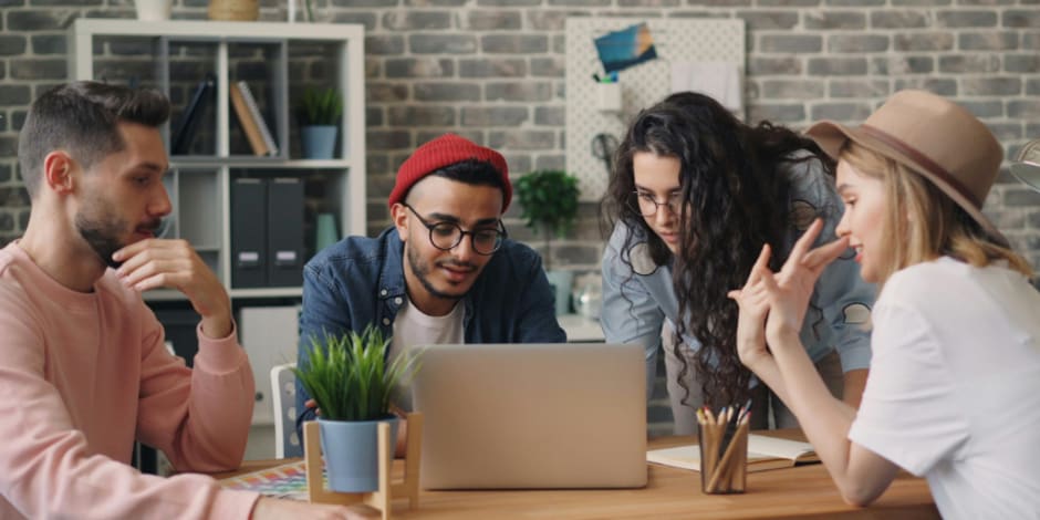 Four people collaborating at office table with laptop, notebooks, and plants