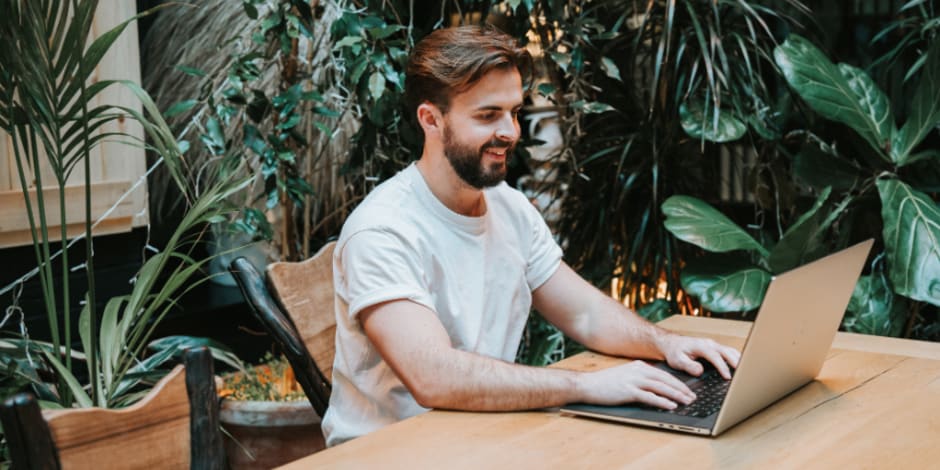 A cheerful man, sitting at a desk, working on his laptop, with plants in the background