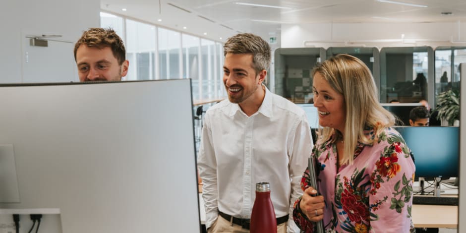 Team collaborating around a computer in an open office, illustrating workplace dynamics or digital strategy planning.